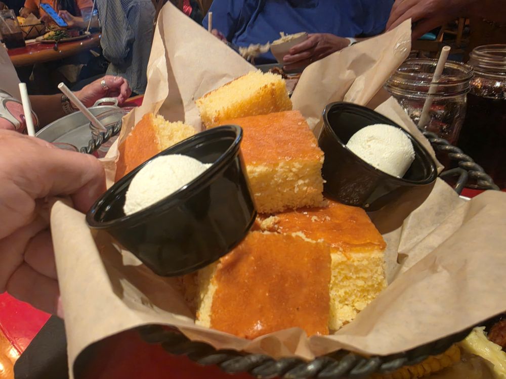 Basket of golden cornbread squares with two black cups of vanilla ice cream on parchment at a crowded restaurant table.