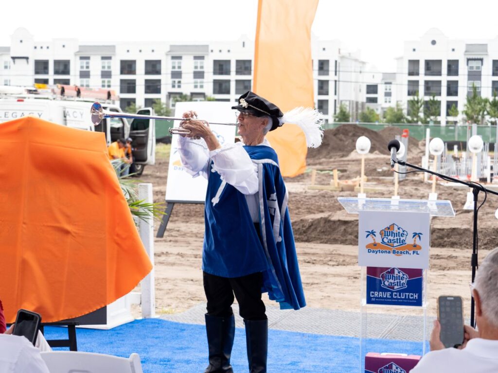 Man in a blue costume and tricorne hat plays a trumpet at an outdoor ceremony on a construction site, with orange drapes and a podium nearby.