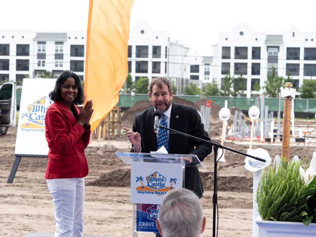 Man in a suit speaks at a clear podium on a construction site, with a woman in red beside him clapping and a White Castle Daytona Beach sign in the background