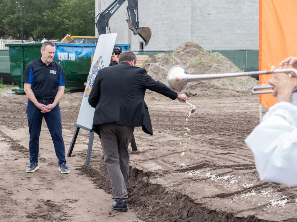 A man in a dark suit pours onions from a ceremonial shovel into a trench at the White Castle Daytona Beach construction site while onlookers and a sign stand nearby.