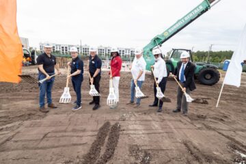 Group of people in hard hats and business casual attire holding shovels at a groundbreaking ceremony on a dirt construction site, with a green crane in the background.