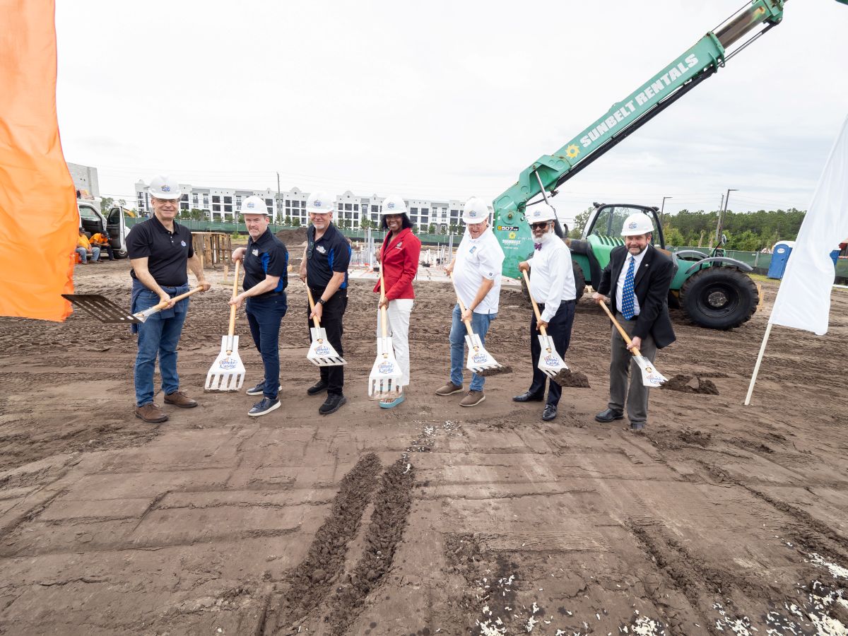 Group of people in hard hats and business casual attire holding shovels at a groundbreaking ceremony on a dirt construction site, with a green crane in the background.