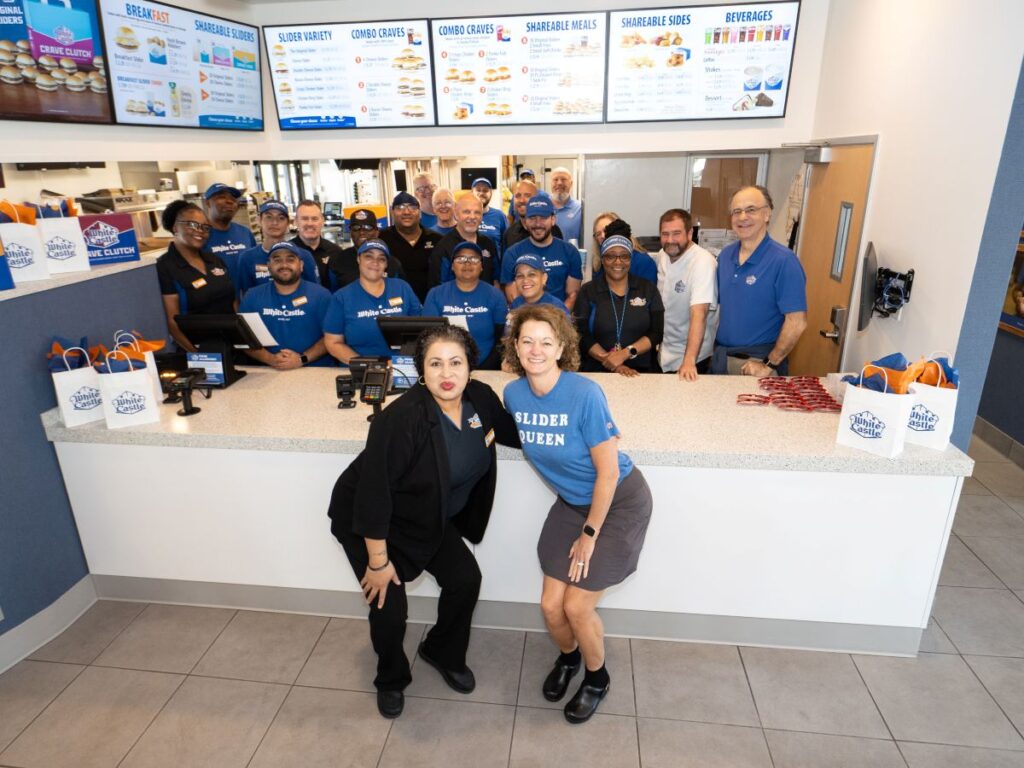 Group of White Castle employees posing behind the counter, two women in front wearing a 'Slider Queen' shirt, smiling at the camera.