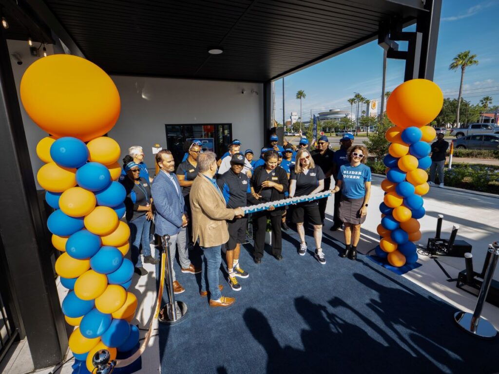 Ribbon-cutting ceremony outside a building with orange and blue balloon columns and a blue carpeted floor, group of people celebrating.