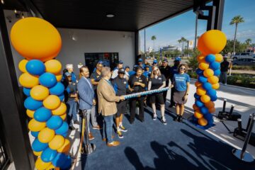 Ribbon-cutting ceremony outside a building with orange and blue balloon columns and a blue carpeted floor, group of people celebrating.