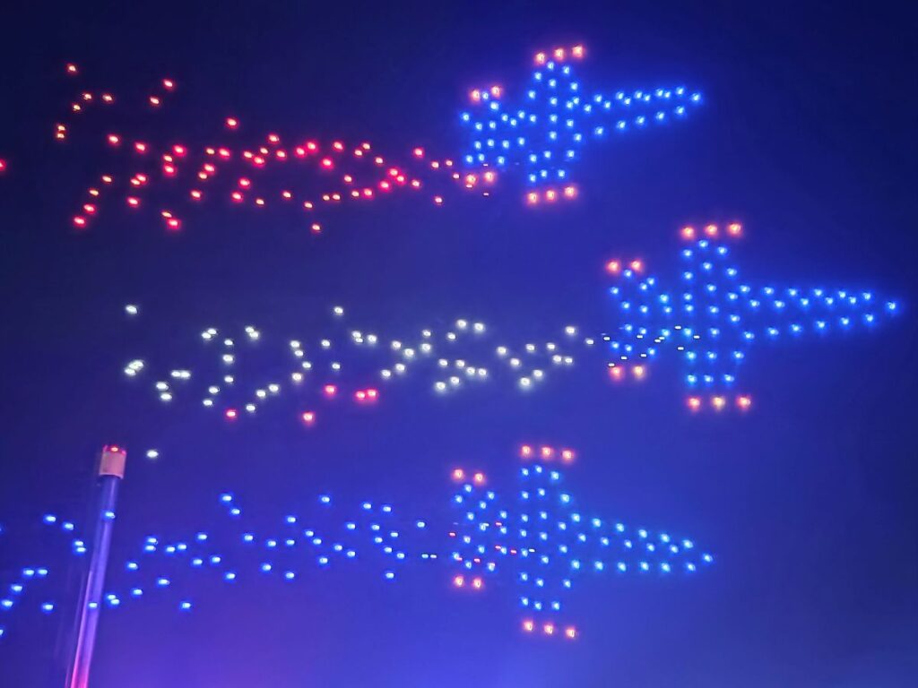 Nighttime drone display showing blue star-shaped airplanes with red and orange accents in the sky.