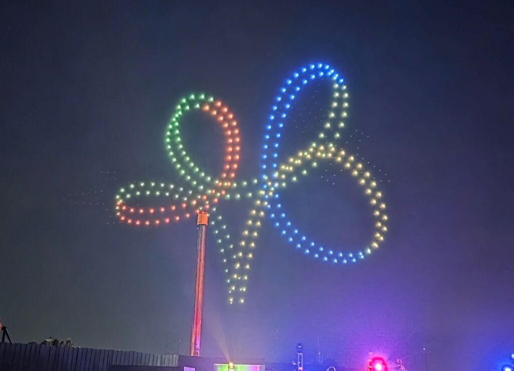 Night scene of a tall pole with a multicolored LED drone light line forming an infinity loop in the sky, green, red, blue, and yellow dots visible in the dark for Summer Nights 2026