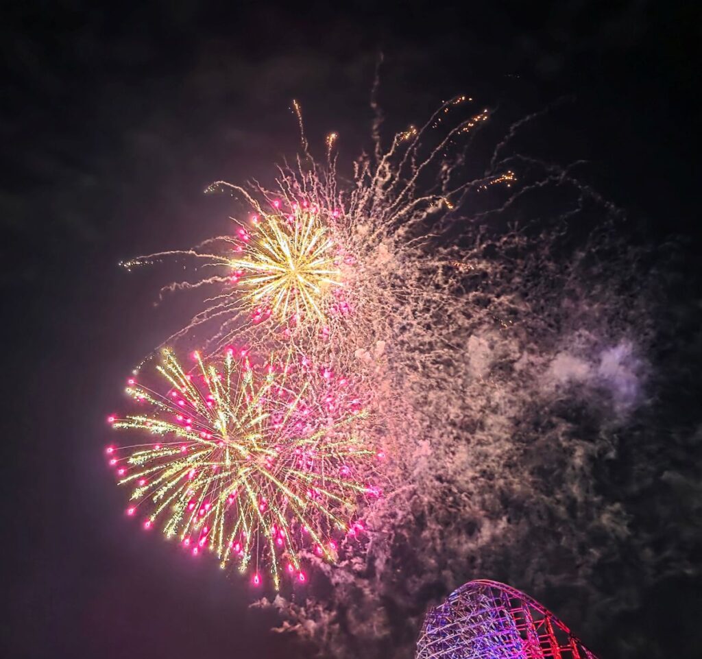 Colorful fireworks exploding in a dark night sky above a roller coaster structure in the lower right corner.