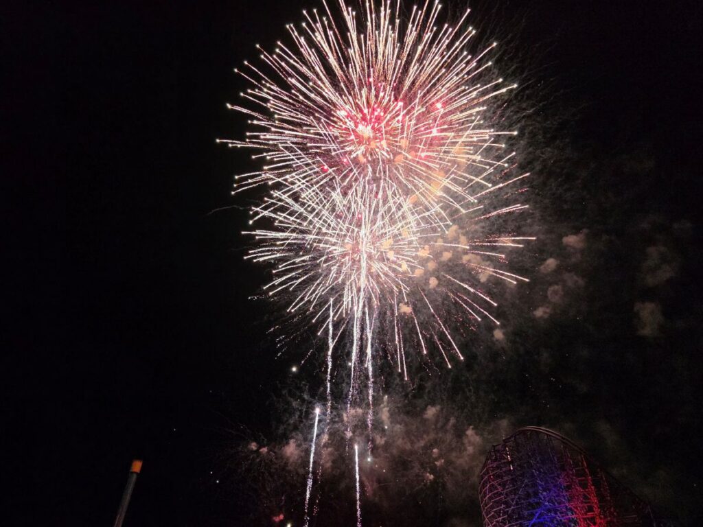 Colorful Summer Nights 2026 fireworks explode in the night sky above a nighttime amusement park with a roller coaster silhouette in the lower right.