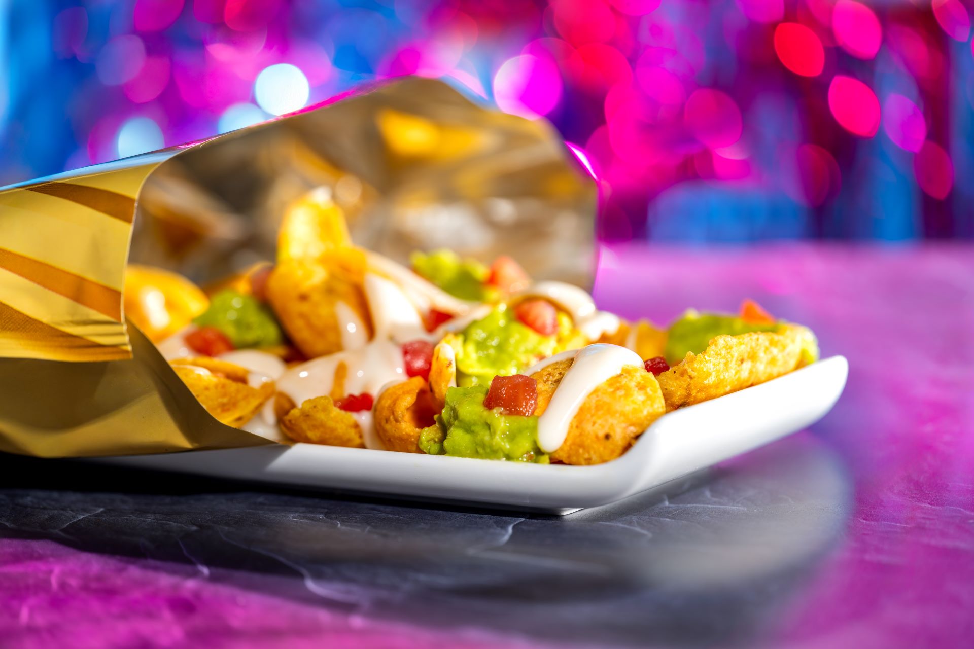 Loaded nachos with cheese, guacamole, and salsa on a white plate, with a bag of tortilla chips nearby and colorful bokeh lights in the background for FØØD by Swedish Chef
