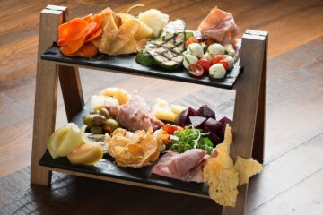 Two-tier wooden charcuterie board with assorted meats, cheeses, olives, fruits, and crackers on black slate platters.