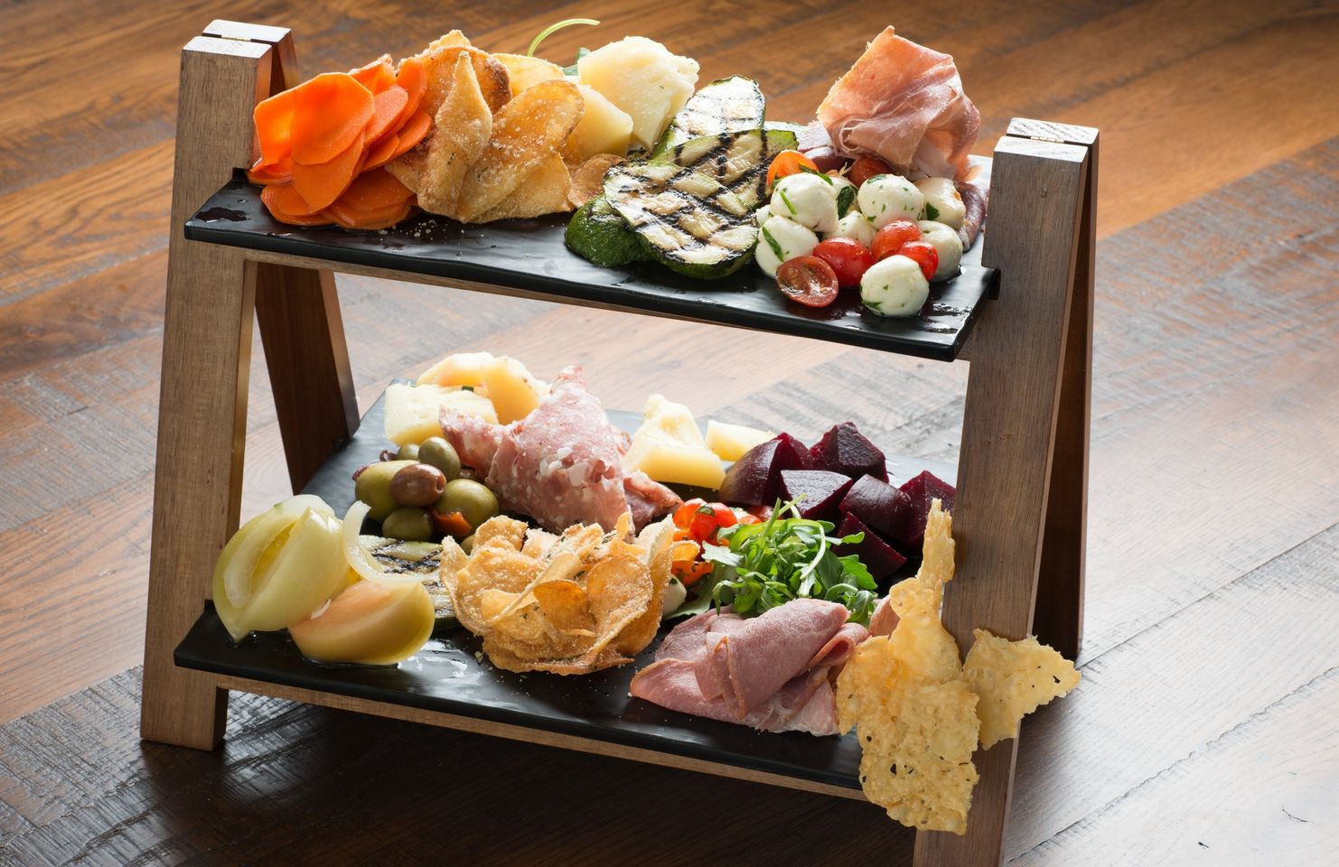 Two-tier wooden charcuterie board with assorted meats, cheeses, olives, fruits, and crackers on black slate platters.