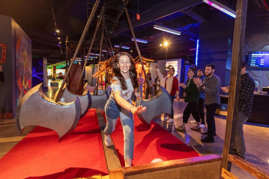Woman in denim runs toward the camera on a suspended axe‑shaped ride as a group of friends cheers in an arcade.