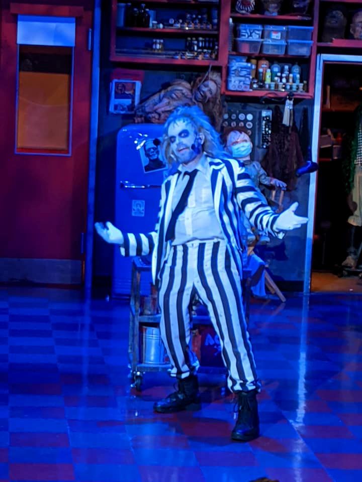 Performer in a black-and-white striped suit with white makeup stands with arms outstretched under blue stage lighting.