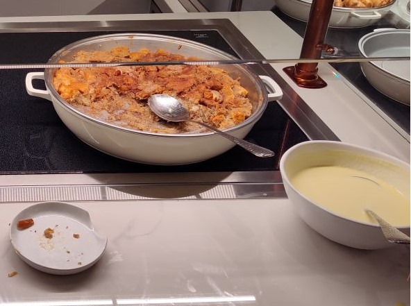White pan on a warming stove filled with crispy fried chicken pieces; a spoon rests inside the pan, with a bowl of pale sauce to the right and a small plate with crumbs in the foreground.