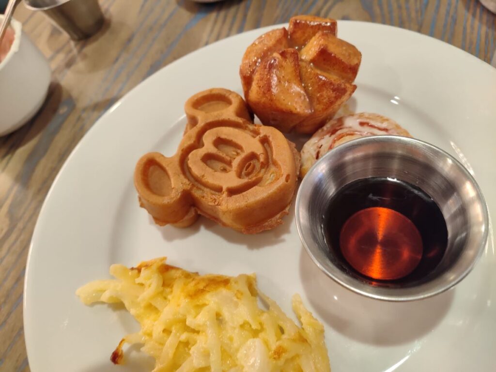 White plate with a Mickey Mouse waffle, a cinnamon roll, hash browns, and a metal cup of dark syrup.
