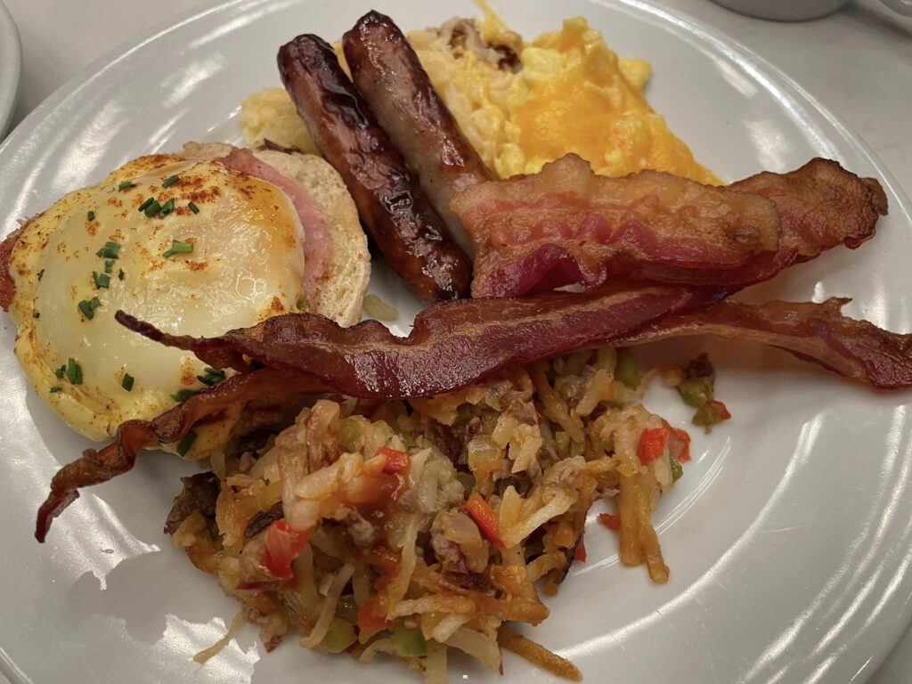 Plate with eggs Benedict on an English muffin, ham, and Hollandaise; crispy bacon, sausage links, scrambled eggs with cheese, and hash browns on the side.