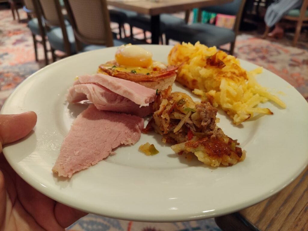 Plate with sliced ham, a hash with onions and peppers, a cheesy potato bake, and a small toasted bread round topped with a fried egg.