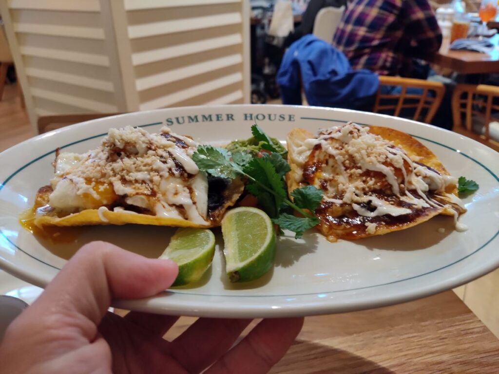 Two tacos with white sauce, cheese, and cilantro on a plate, plus lime wedges, in a casual restaurant setting labeled Summer House.