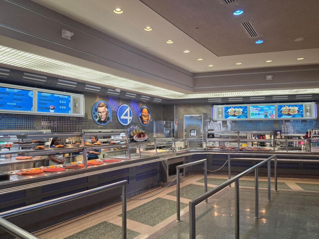 Cafeteria-style serving line with glass barriers, digital menu boards, and workers behind the counter serving plates of food in a blue-tiled dining hall.