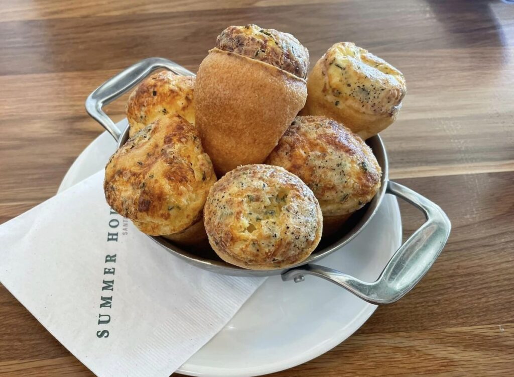 Small savory cheese and herb rolls piled in a metal skillet over a white plate, on a wooden table; napkin with 'SUMMER HOME' partially visible nearby.