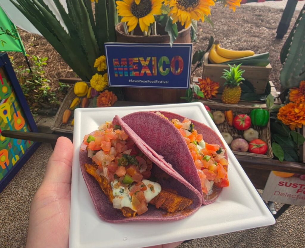 Blue corn tacos filled with meat and pico de gallo on a white plate, held in front of a Mexico festival display with sunflowers and produce in the background.