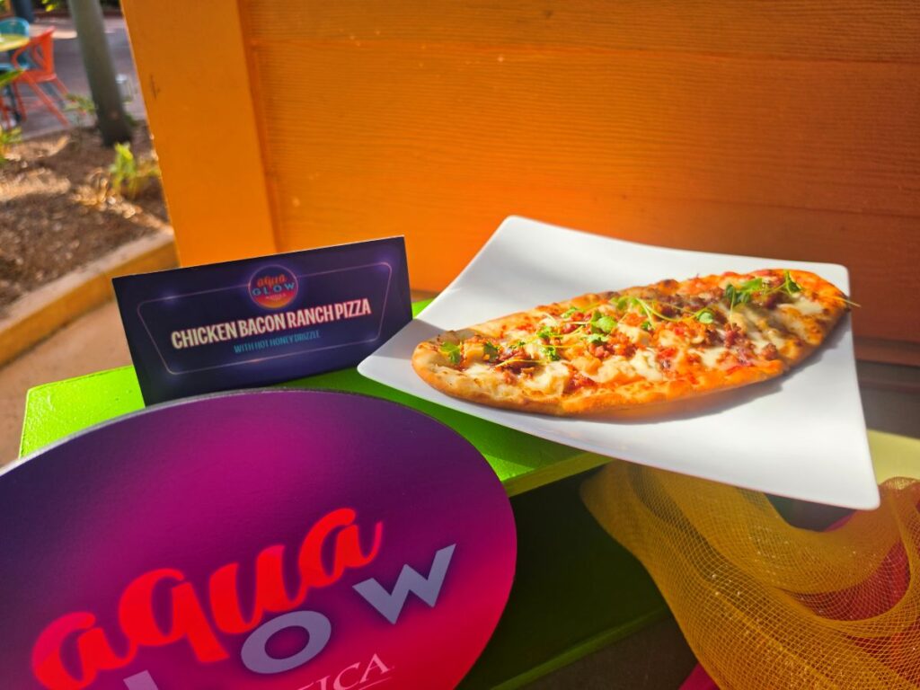 A rectangular pizza topped with cheese and toppings on a white plate outdoors, with a bright orange wall in the background.