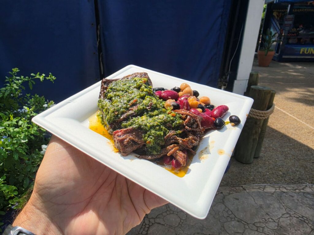 White square plate held in a hand, featuring a herb-topped steak with green sauce and a side of mixed olives and colorful beans; outdoor market setting in the background at Seven Seas Food festival