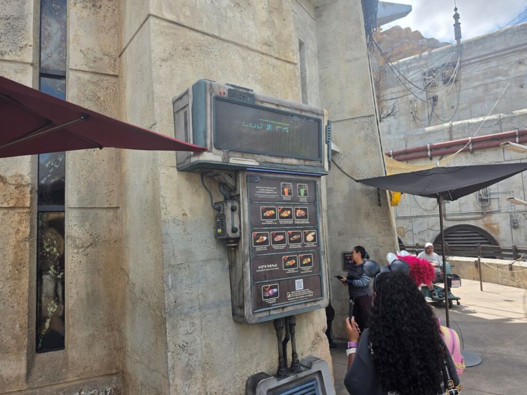 Outdoor stone-wall display with a large electronic menu board on a futuristic metal panel; people stand nearby under umbrellas in a sunny courtyard.