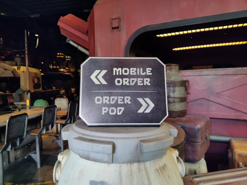 Informational sign reading 'MOBILE ORDER' with arrows, atop stacked beige containers in a sci-fi themed dining area.
