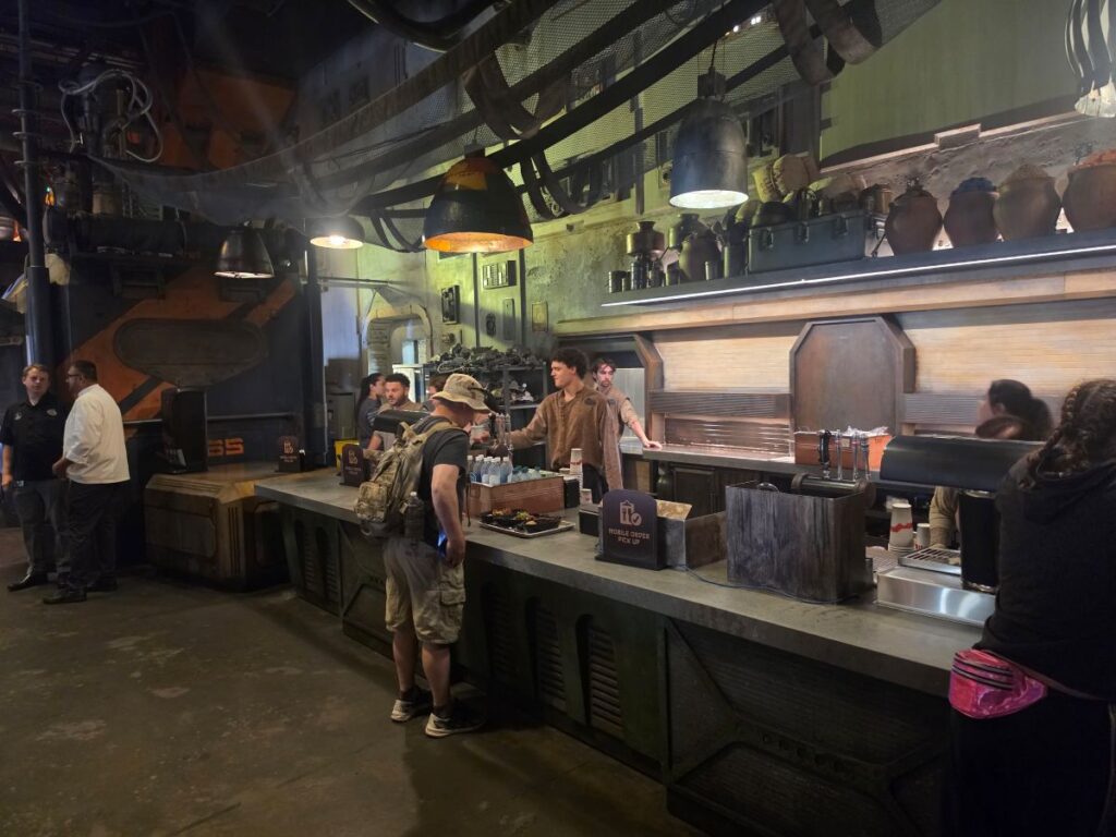 Industrial-style bar or cafe with a long counter, customers ordering, and staff serving under large hanging lights.