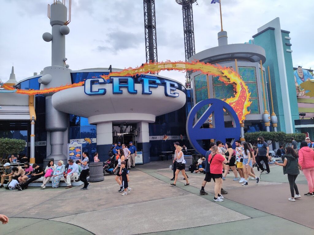 Cafe 4 entrance at a theme park, with a large circular blue 4‑shaped sculpture and visitors walking by.