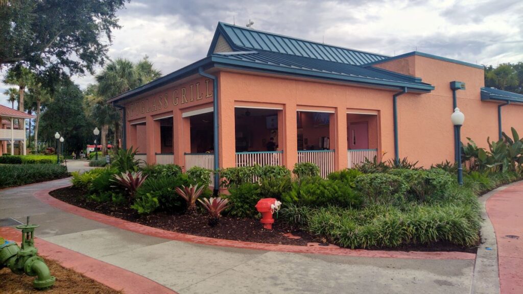 Peach-colored restaurant with blue trim and an open-front dining area, surrounded by tropical plants and a curved walkway.