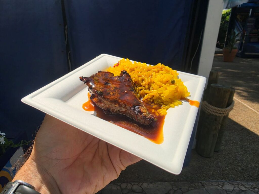 Plate held outdoors with glazed grilled lamb chop and yellow rice on a white disposable plate.