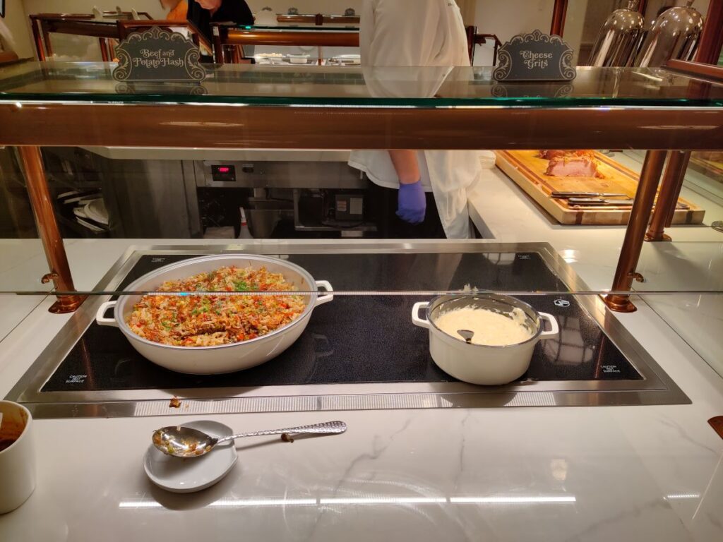 Buffet counter with a large white pot of beef and potato hash on a black induction surface, labeled nearby, and a pot of cheese grits to the side; chef in white uniform in background wearing a purple glove.