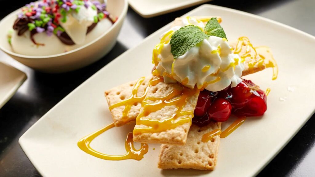 Hollywood & Vine lunch dessert plate with graham crackers, whipped cream, caramel drizzle, and cherry compote, garnished with a mint leaf.