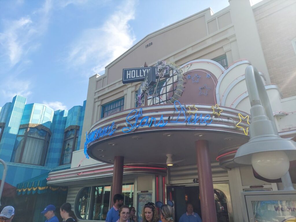 Exterior of a retro-style venue with a neon marquee and star decorations; people stand outside under a blue sky.