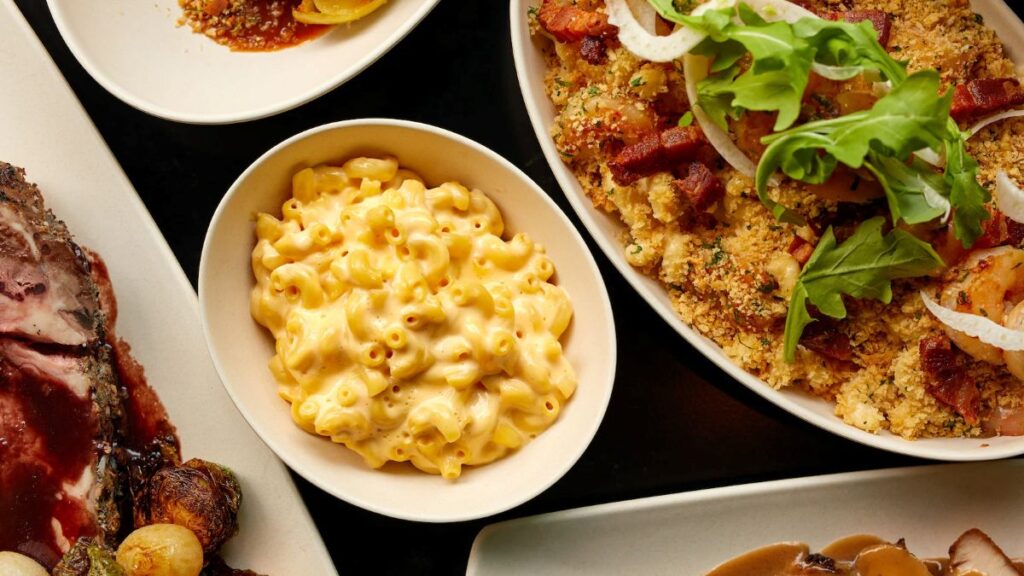 Creamy macaroni and cheese in a white bowl, with roasted meat on the left and a crumb-topped dish with greens on the right