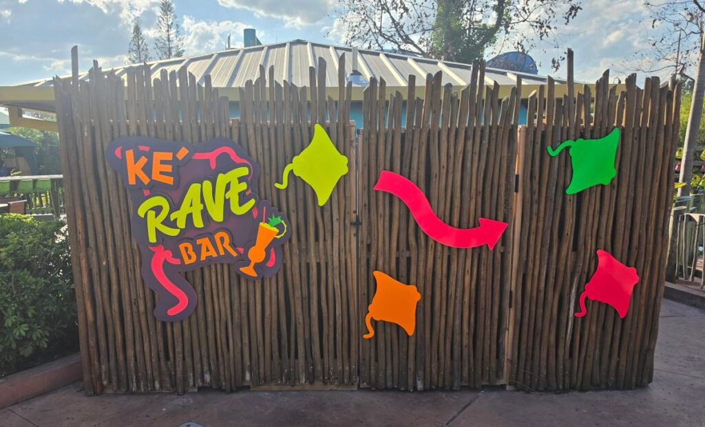 Colorful wooden fence with a bright sign reading 'KE RAVE BAR' and playful neon shapes decorating a party venue exterior.