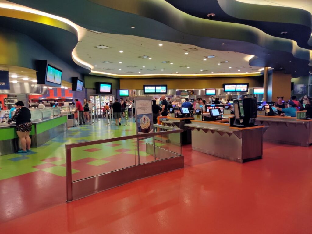 Colorful food court with curved ceiling, service counters, and customers ordering at kiosks and counters.