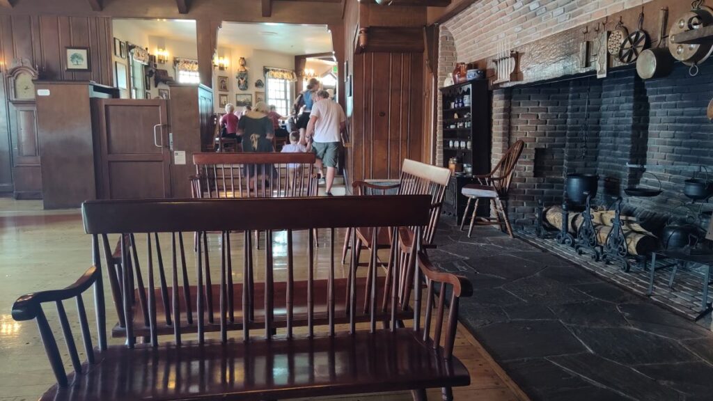 Rustic dining hall with dark wooden benches in the foreground and a group of people gathered near the back, brick fireplace with hanging pots on the right. photo for Liberty Tree Tavern review