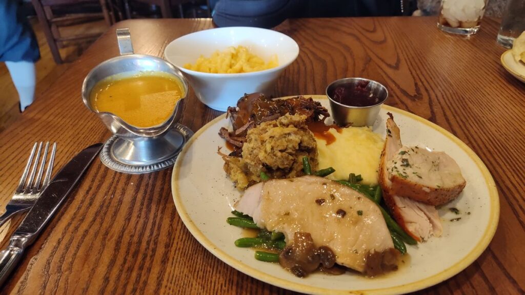 Plate with sliced turkey, stuffing, mashed potatoes, green beans, cranberry sauce, and a slice of herb-roasted meat on a wooden table; a gravy boat and side dish nearby.