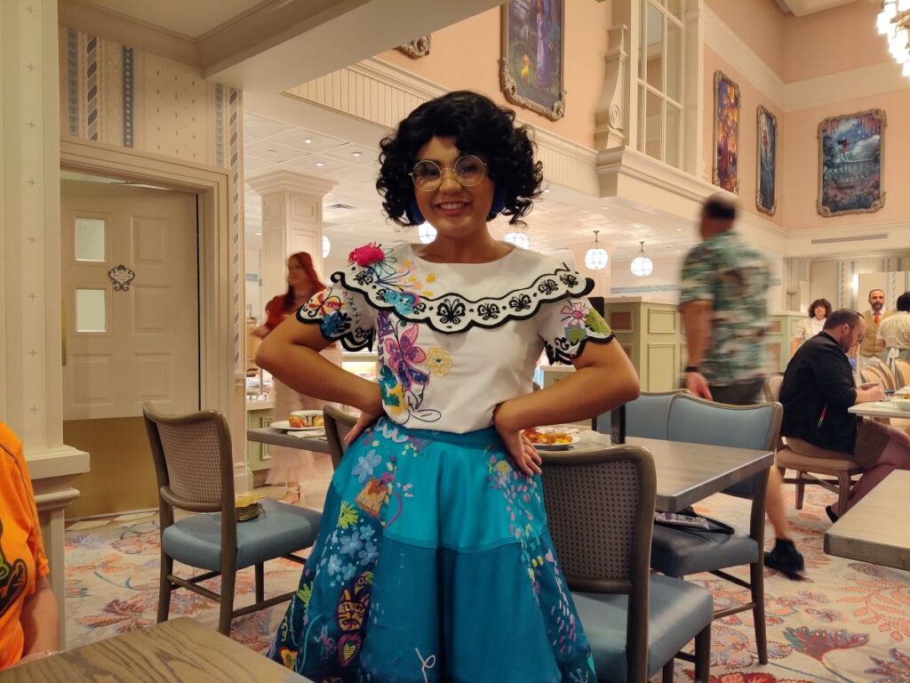 Smiling woman, Mirabel, in a white embroidered top and bright blue floral skirt stands with hands on hips in a bright dining hall.