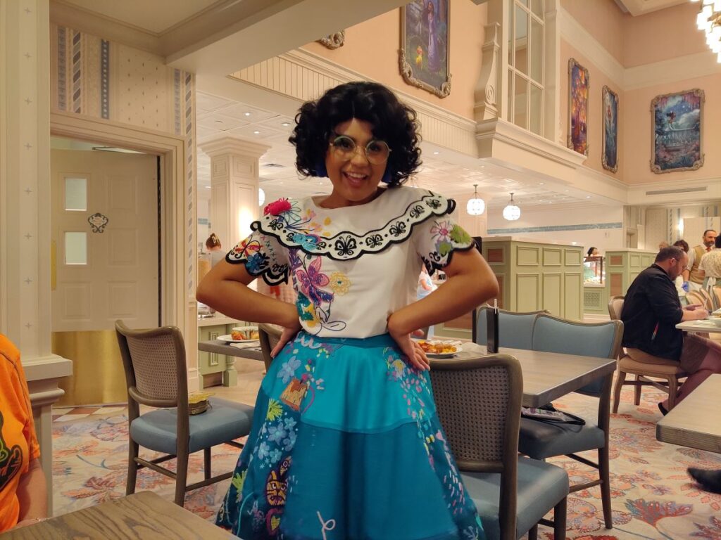 Person wearing a white embroidered blouse and bright blue floral skirt stands with hands on hips in a pastel-lit dining room, smiling at the camera.