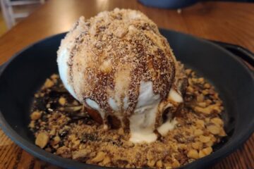 Skillet dessert with a large scoop of vanilla ice cream, caramel drizzle, and a crumbly nutty crust around the base in a black cast-iron pan