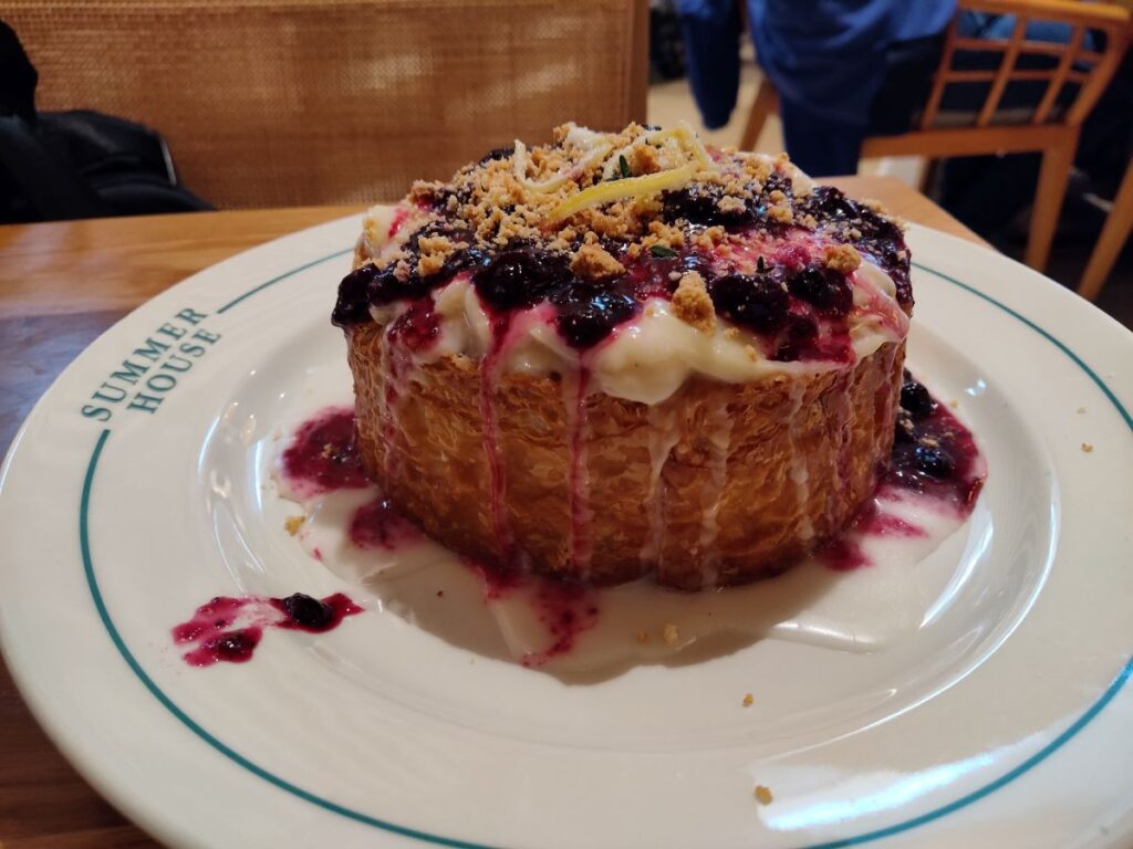 Round cake with white icing and blueberry sauce, topped with crumble, on a plate labeled 'Summer House'.