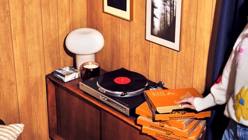 Turntable on a wood-paneled cabinet with a red-centered vinyl, a lamp, candle, and a stack of orange boxes nearby. IMage is for Blaze Pizza Delivery Offer