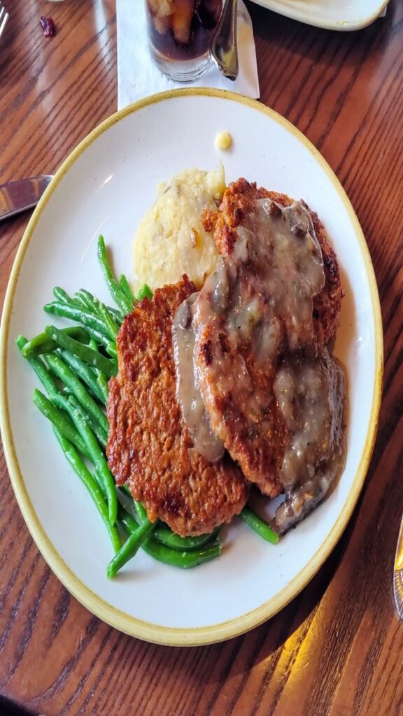 Fried chicken cutlets with brown gravy, mashed potatoes, and green beans on a white plate with a wooden table.
