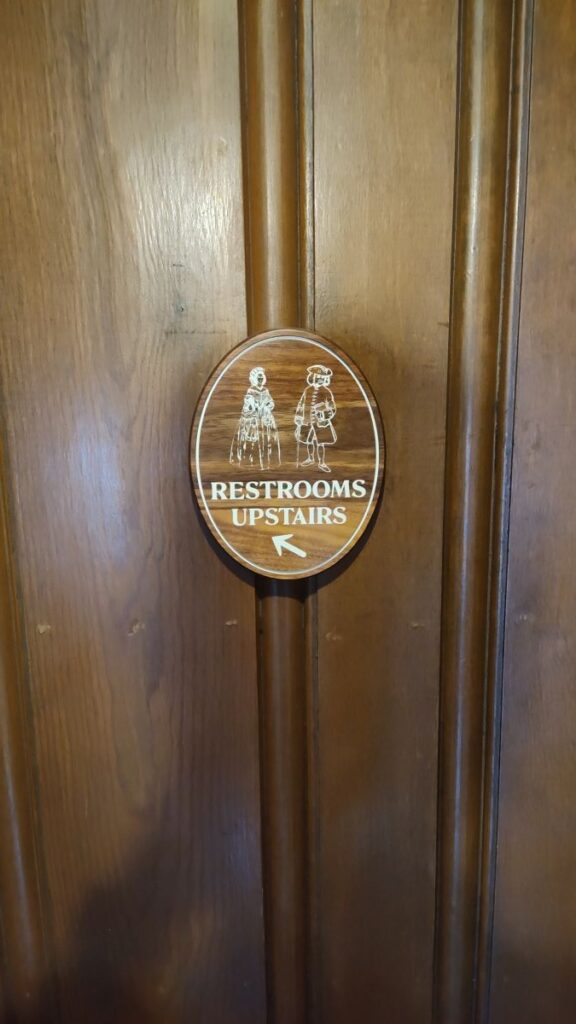 Round wooden sign on a door reading 'Restrooms Upstairs' with silhouettes of a man and a woman and a leftward arrow.