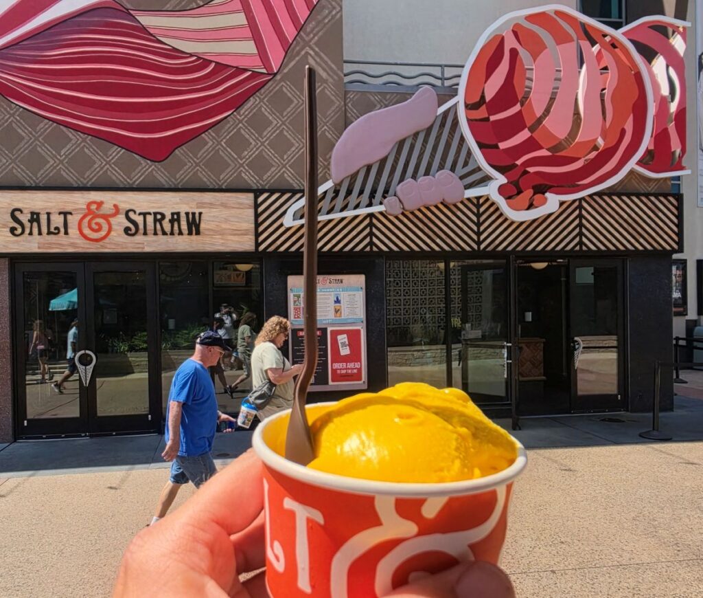 Close-up of a bright yellow ice cream scoop in an orange Salt & Straw cup, held up in front of a mural storefront.